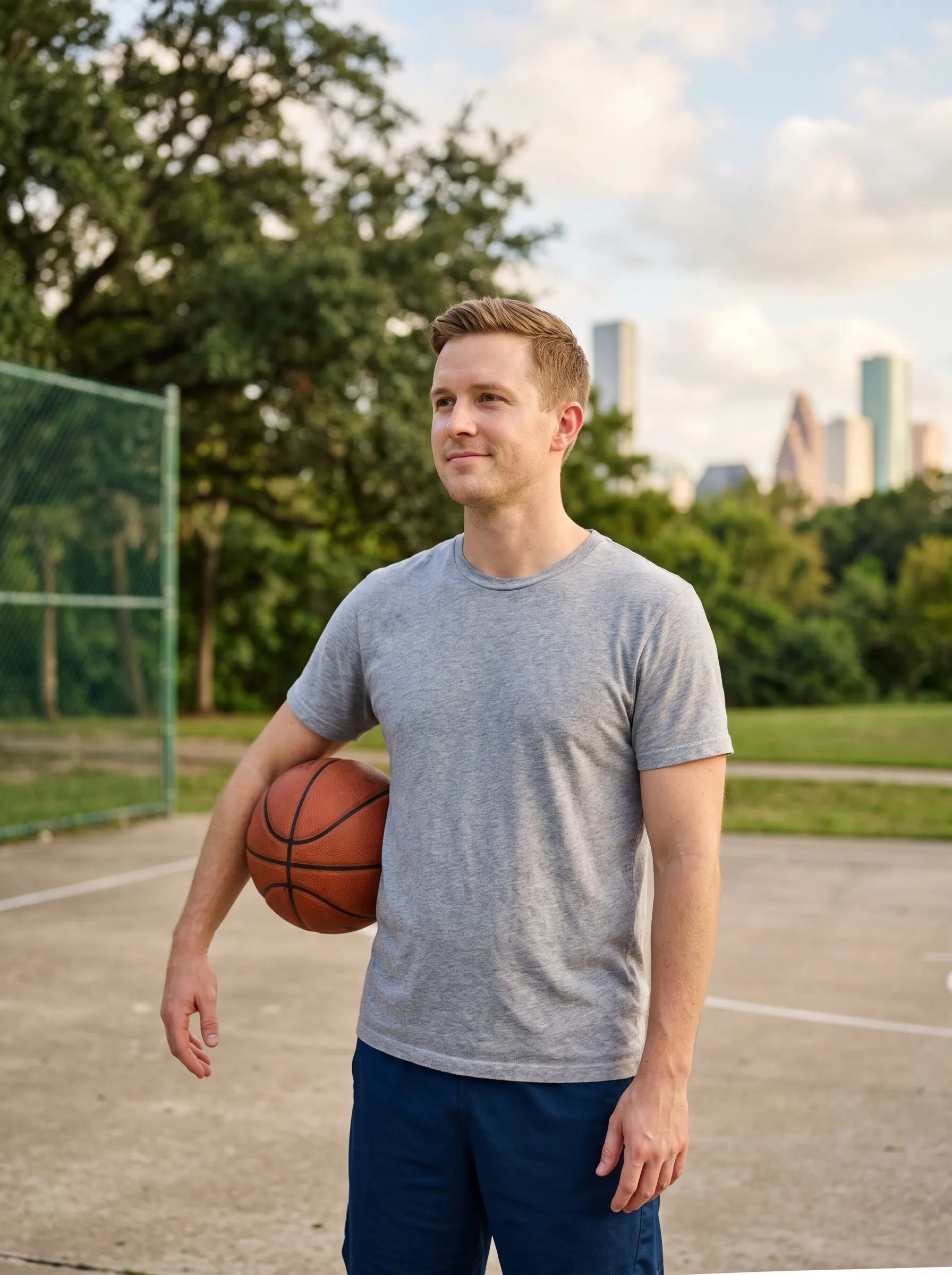 A man in his early thirties holding a basketball after a relaxed shoot-around at a leafy Houston park court, calm and content, the downtown skyline soft in the distance.