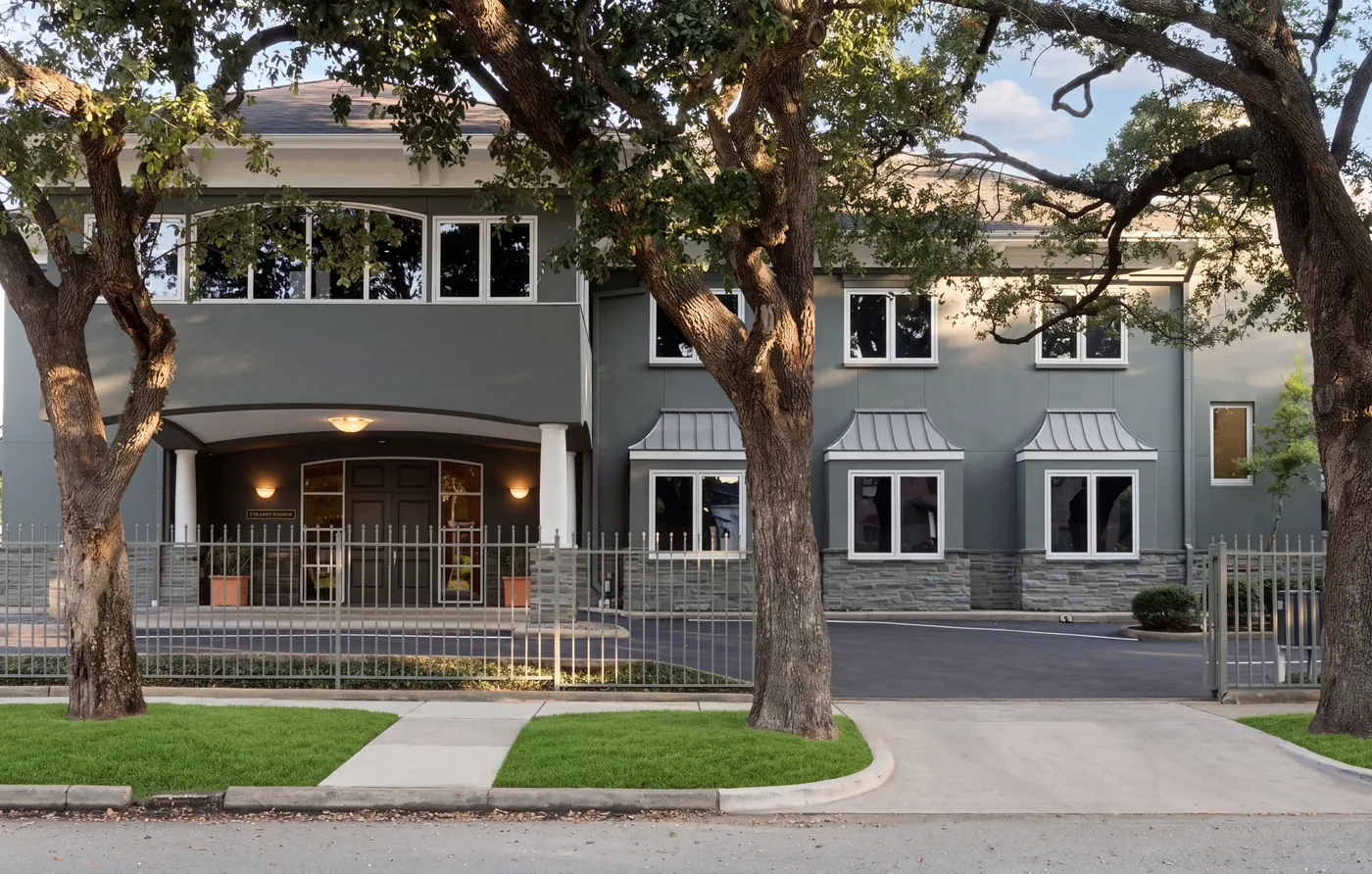 The Lovett Center exterior at 900 Lovett Boulevard: a two-story stucco building with an arched portico, set back from the street under mature oaks in Montrose.