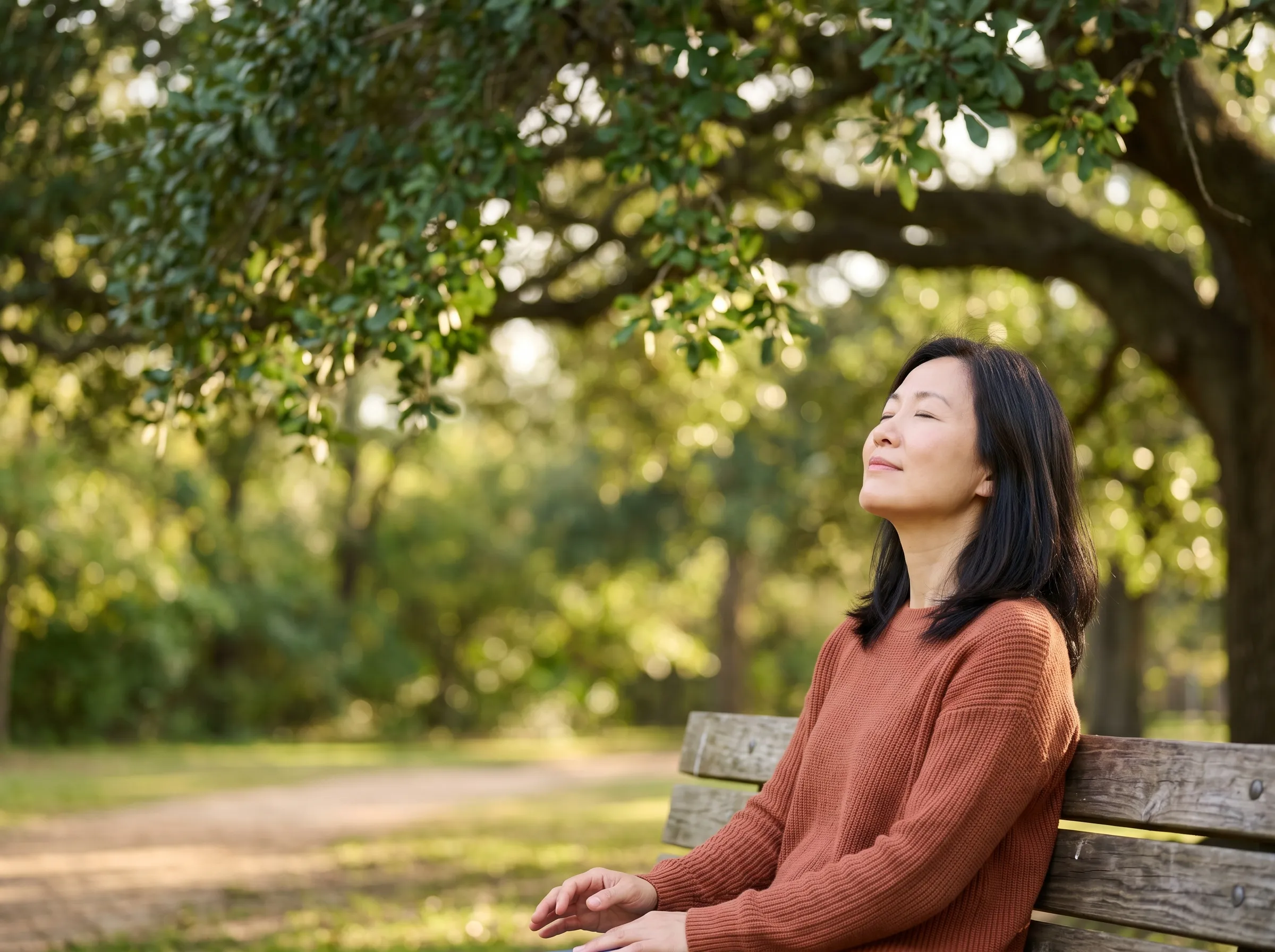 A woman sitting on a park bench under Houston live oaks, face turned to soft sunlight, calm and at ease.